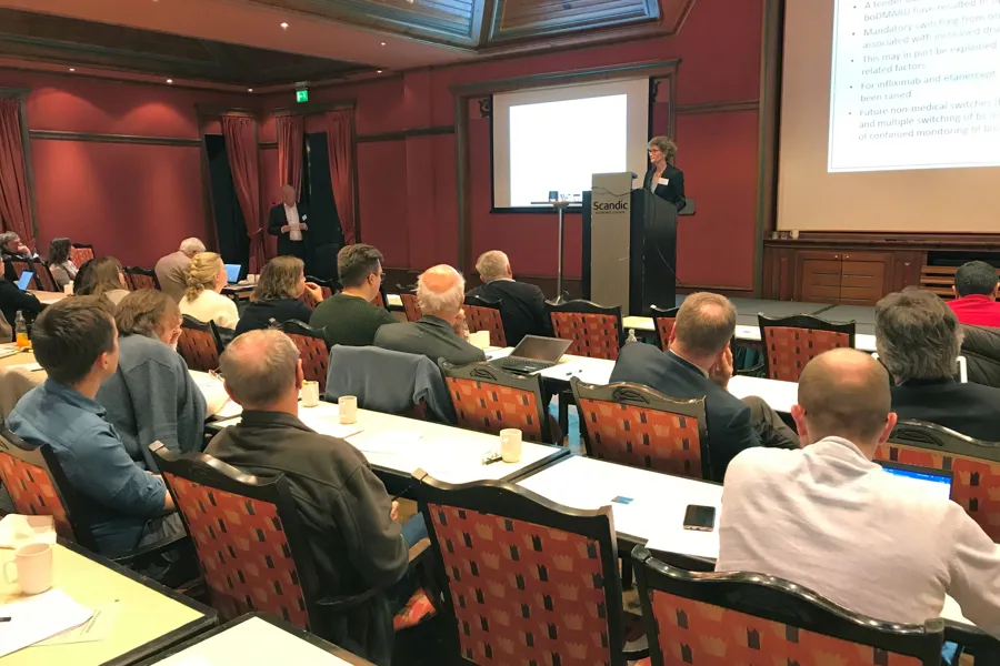 En rekke personer sitter i en konferansesal mens en åerspn står på en talerstol og snakker til forsamlingen.